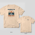 Back and front of an ivory T-shirt: back shows a kayak angler in an arched marsh sunrise scene with Paddle In Fish Out; front has a small kayak logo