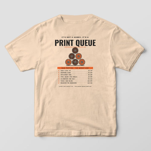 Back print on an Ivory Comfort Colors 1717 tee: "It's Not a Hobby, It's a Print Queue" over a pyramid of six orange and charcoal spools, with a seven-item Now Printing queue list and print times