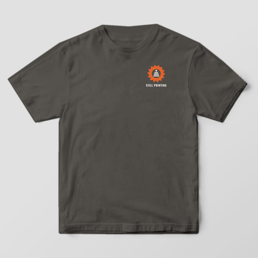Front of the Pepper Comfort Colors 1717 tee showing the left-chest print: Small left-chest orange gear badge with a nozzle stacking layers and "Still Printing"