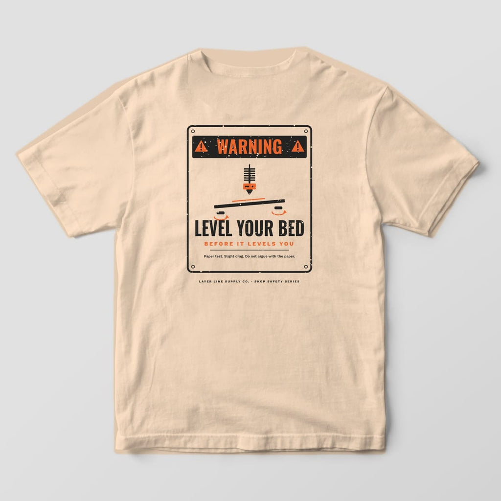 Back print on an Ivory Comfort Colors 1717 tee: Distressed warning sign: "Warning" header, nozzle over a tilted print bed with knobs, "Level Your Bed, Before It Levels You" and "Paper test. Slight drag. Do not argue with the paper."