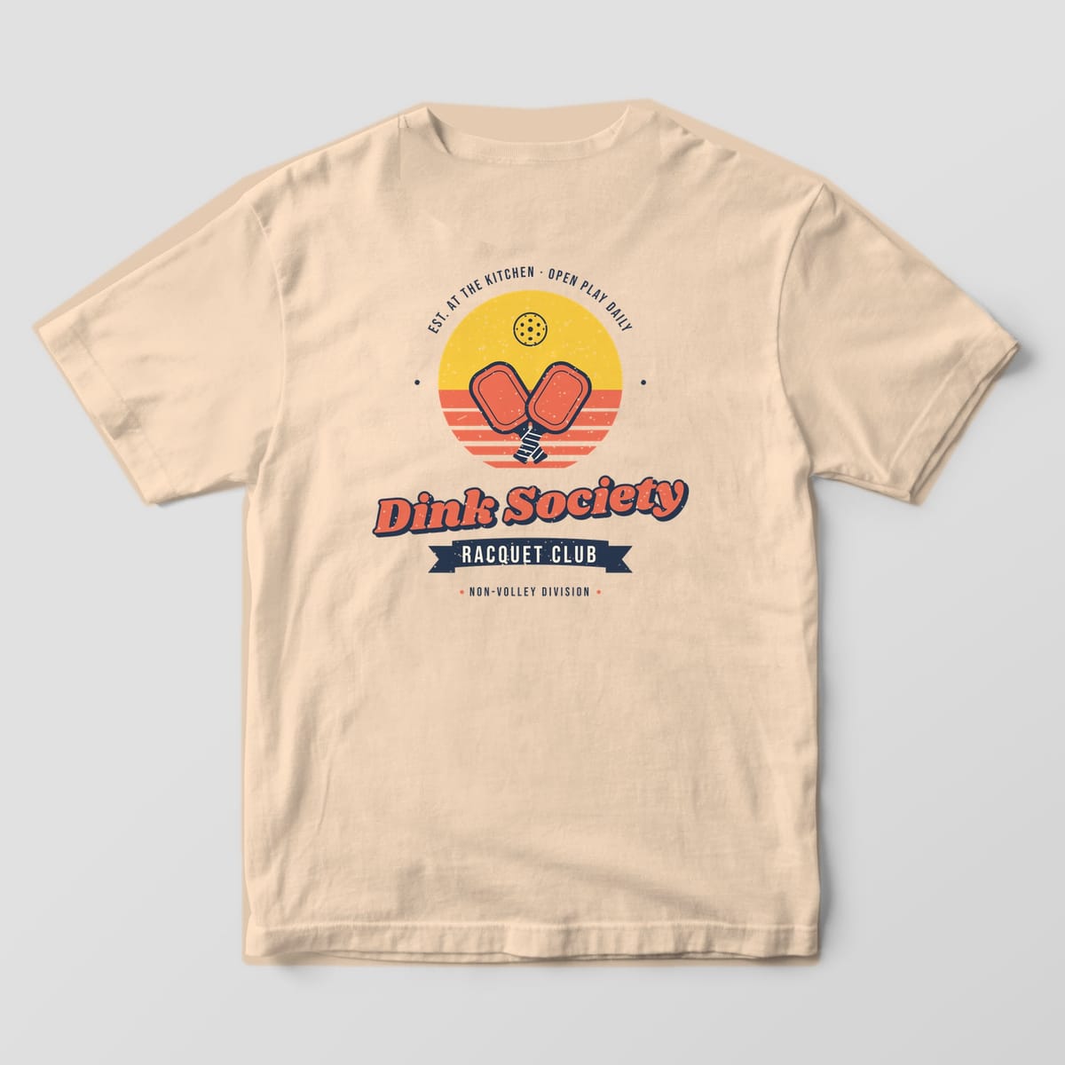 Back print on an Ivory Comfort Colors 1717 tee: Dink Society Racquet Club badge with crossed coral paddles over a striped yellow sunset