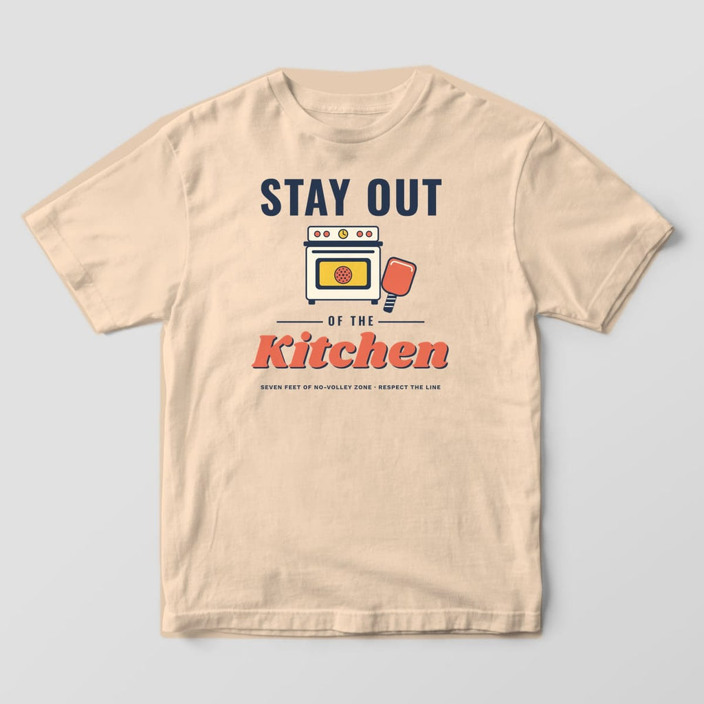Front of the Ivory Comfort Colors 1717 tee showing the full-front print: Full-front print: "Stay Out" in bold navy, a cream retro stove with a pickleball in the oven and a coral paddle, then "of the Kitchen" in puffy coral script and "Seven feet...