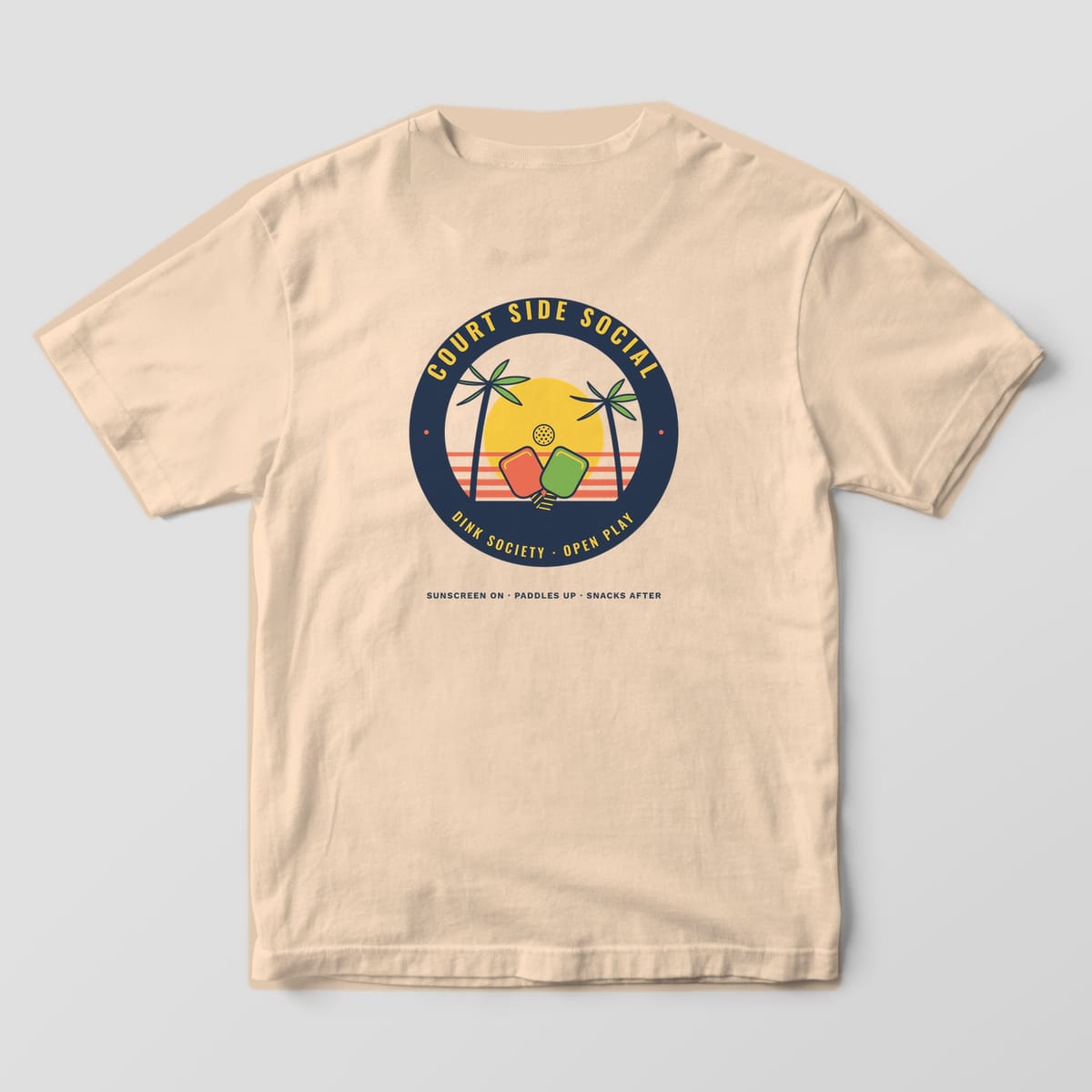 Back print on an Ivory Comfort Colors 1717 tee: Round tropical badge with palm trees, crossed paddles and a yellow sun ringed by Court Side Social