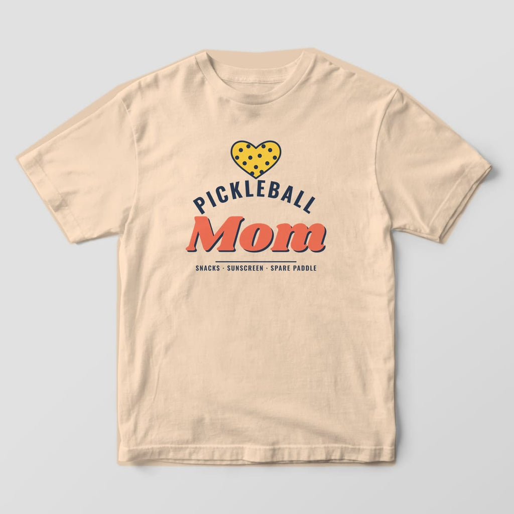 Front of the Ivory Comfort Colors 1717 tee showing the full-front print: Full-front print: a yellow pickleball heart over arched navy "Pickleball", a big coral "Mom" in puffy script and "Snacks, Sunscreen, Spare Paddle"