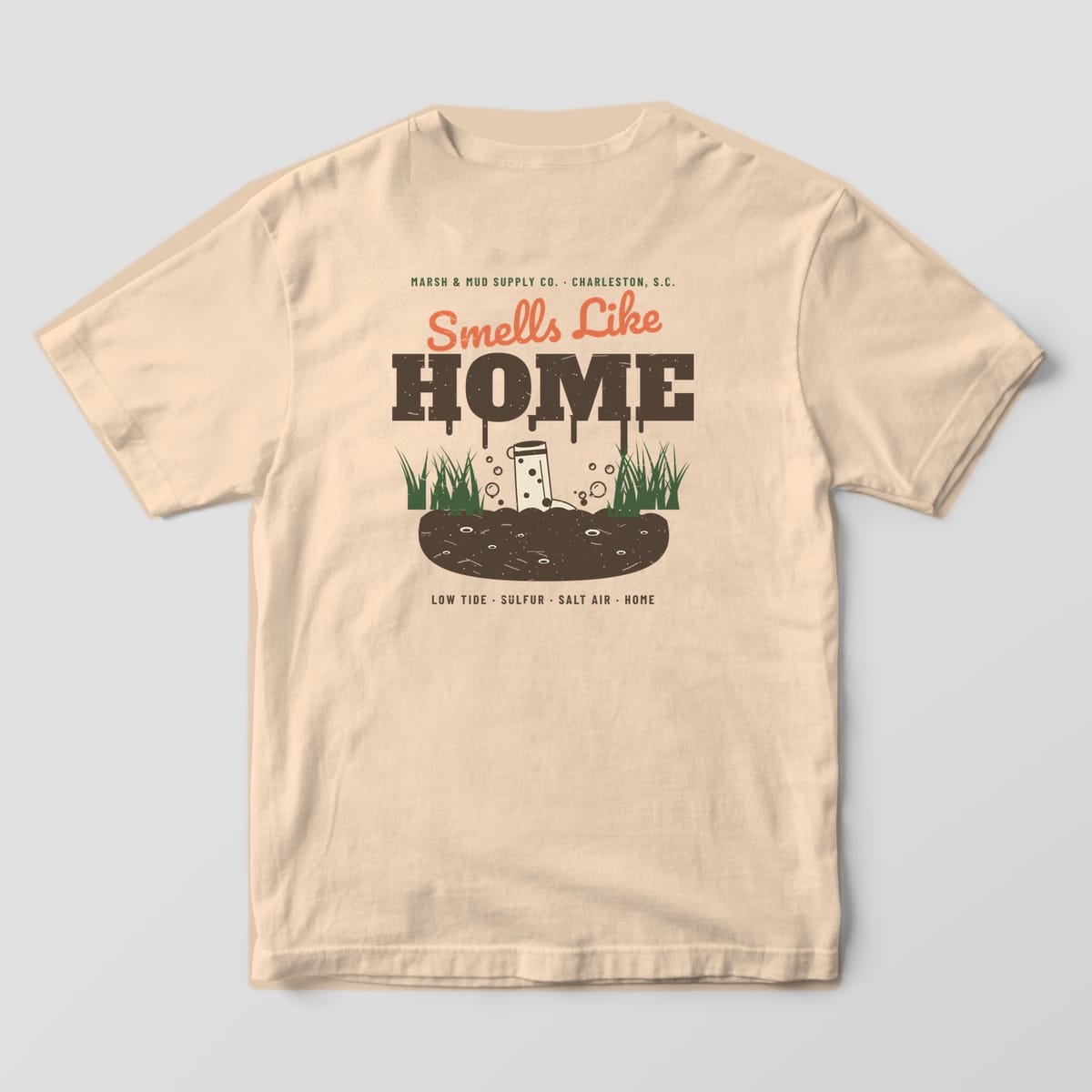 Back of an ivory Comfort Colors 1717 tee with dripping HOME lettering, a rubber boot in mud and green marsh grass