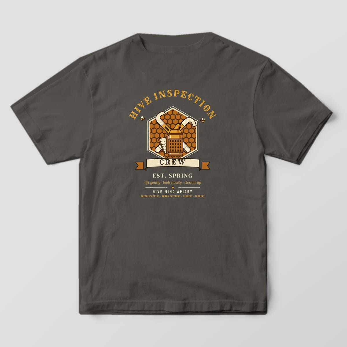 Back of a pepper Comfort Colors 1717 T-shirt printed with a crest of crossed hive tools and a bee smoker on a honeycomb hexagon titled Hive Inspection Crew.