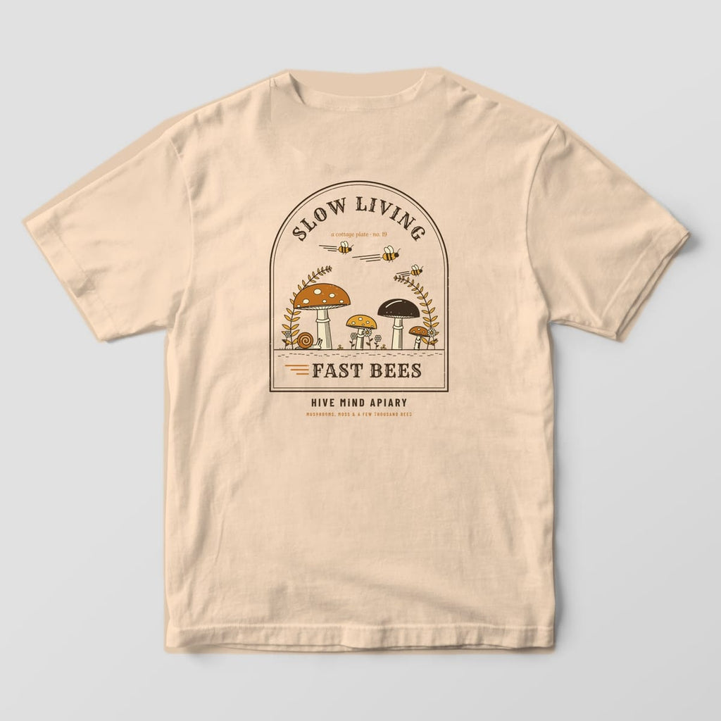 Back of an ivory Comfort Colors 1717 T-shirt printed with an arch-framed cottage scene of mushrooms, ferns and a snail with bees zooming overhead, titled Slow Living Fast Bees.