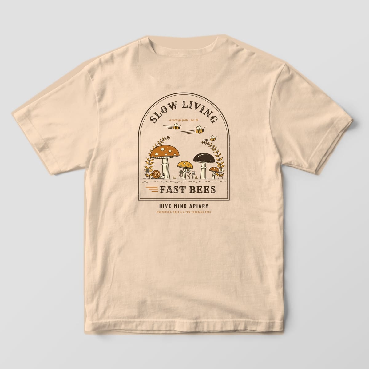 Back of an ivory Comfort Colors 1717 T-shirt printed with an arch-framed cottage scene of mushrooms, ferns and a snail with bees zooming overhead, titled Slow Living Fast Bees.