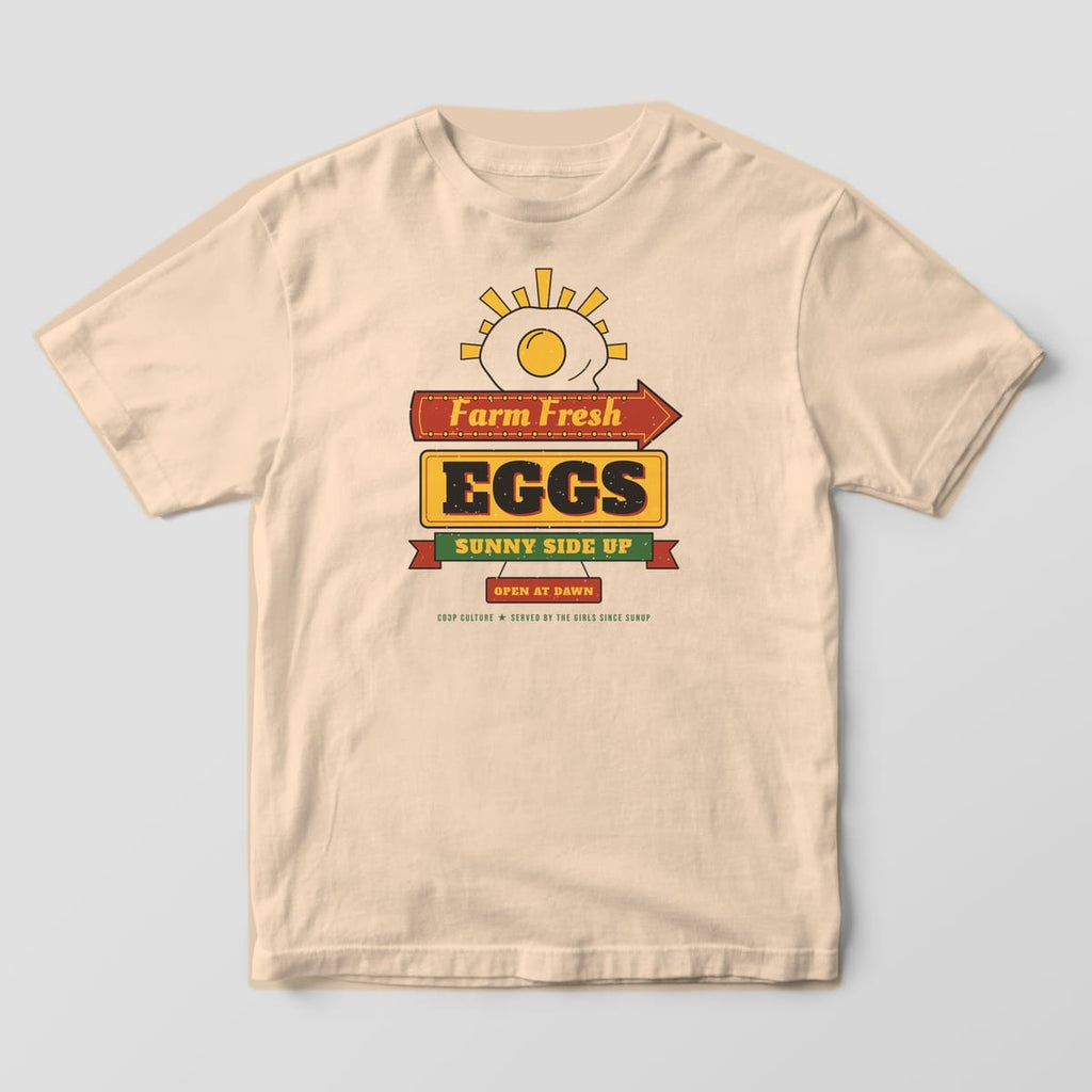 Front of an ivory Comfort Colors tee with a full-front retro diner sign reading Farm Fresh Eggs Sunny Side Up under a fried egg sunrise.