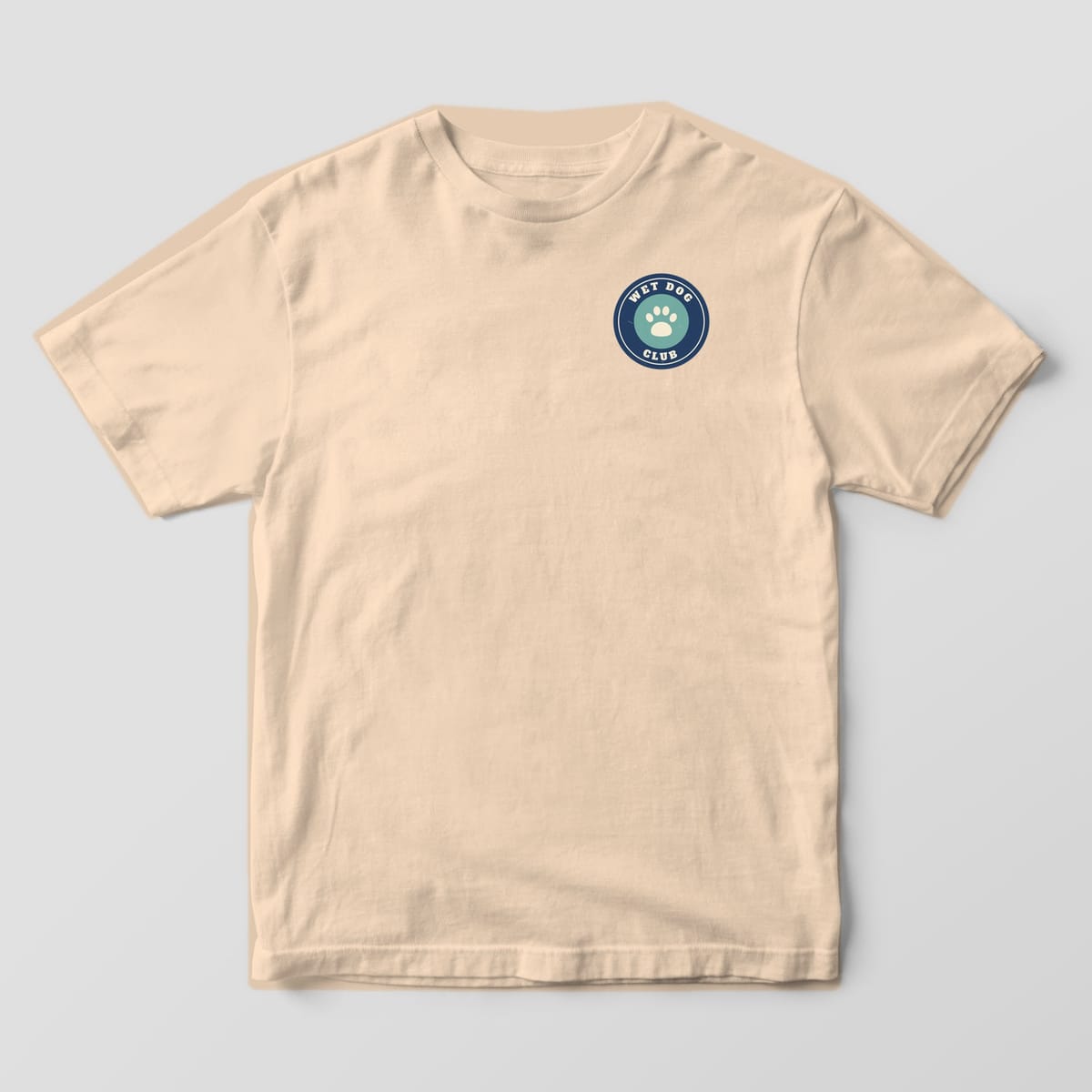 Front of an ivory Comfort Colors tee with a small navy and teal Wet Dog Club paw badge