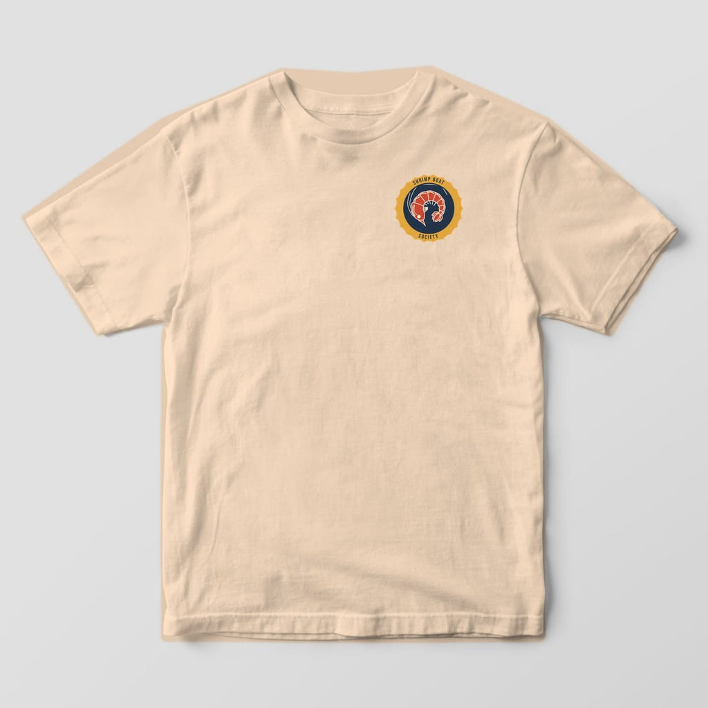 Front of an Ivory Comfort Colors 1717 tee with a small scalloped gold and navy shrimp badge on the left chest.