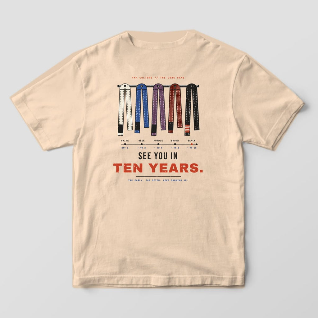 Back of an ivory Comfort Colors 1717 tee with white, blue, purple, brown and black belts on a peg rail and a rank timeline.