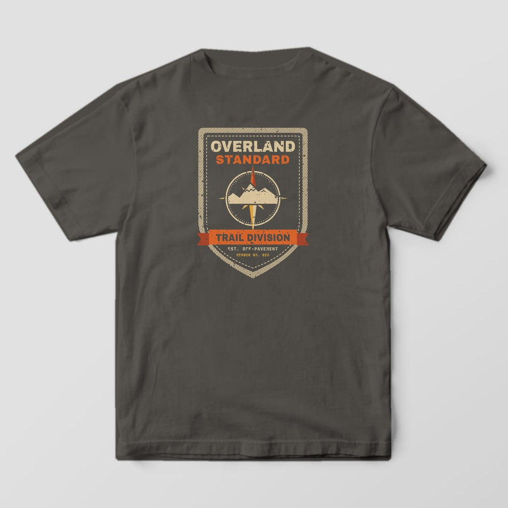 Back of a pepper Comfort Colors 1717 T-shirt showing a vintage stitched shield patch with a compass rose and mountains, a Trail Division ribbon and the words Overland Standard