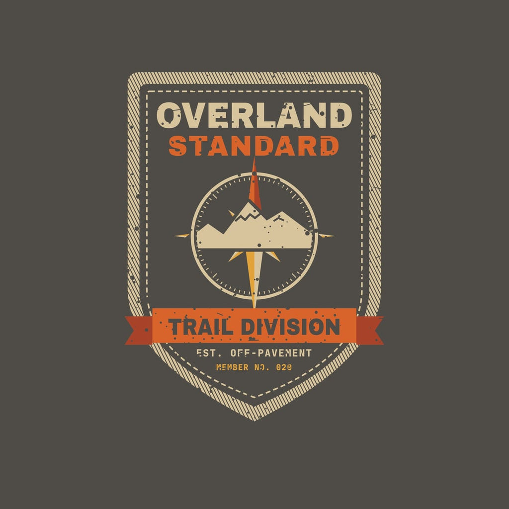 Close-up of the back print artwork on a pepper background: a vintage stitched shield patch with a compass rose and mountains, a Trail Division ribbon and the words Overland Standard