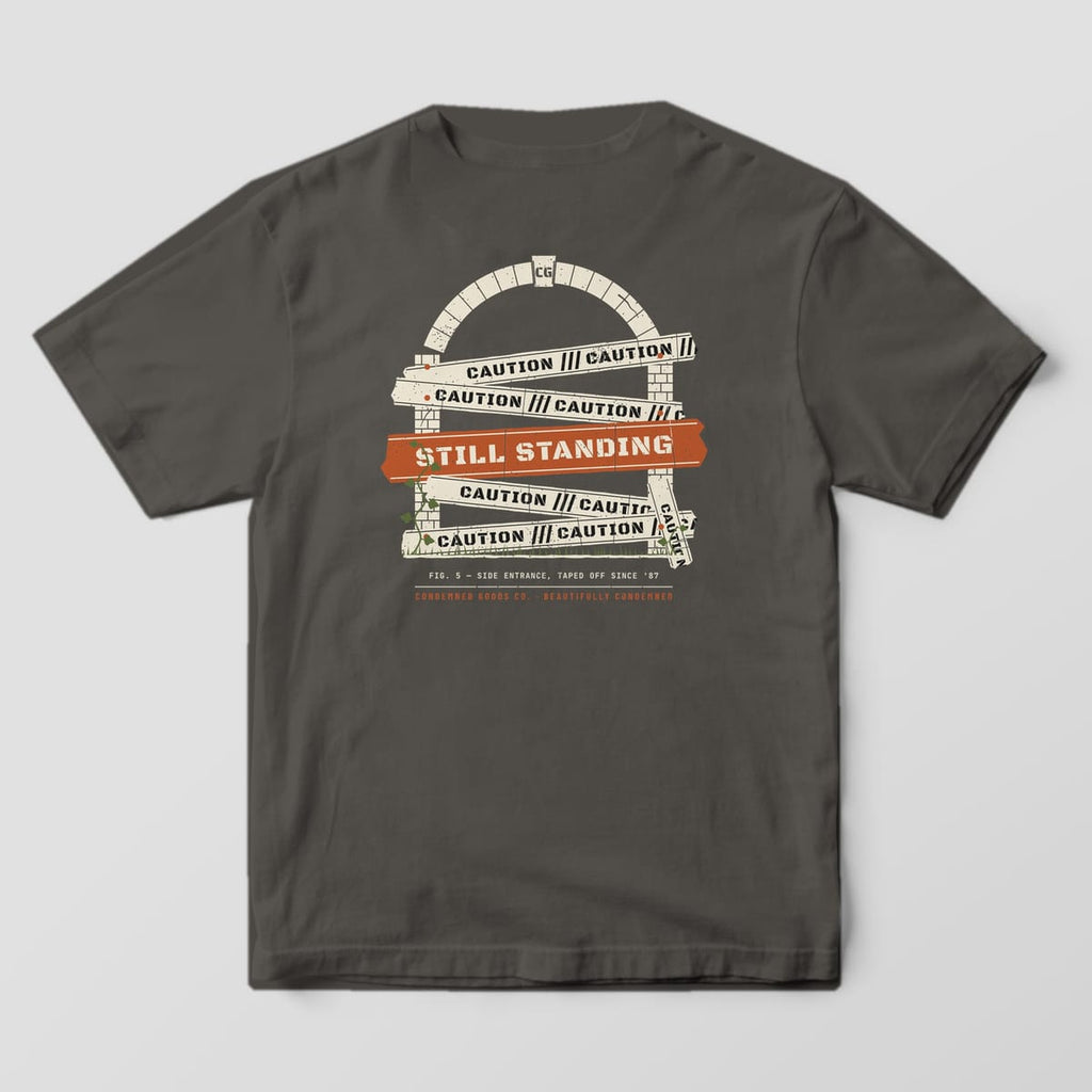 Back of a pepper Comfort Colors 1717 tee with cream caution tape strips across a brick arch and a rust Still Standing band.
