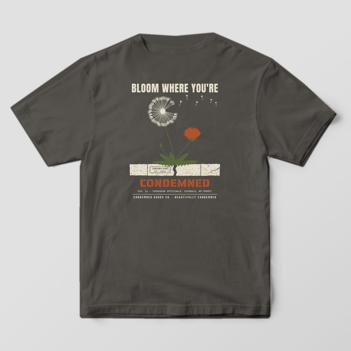 Back of a pepper Comfort Colors 1717 tee with a dandelion growing from a cracked sidewalk slab and the words Bloom Where You're Condemned.