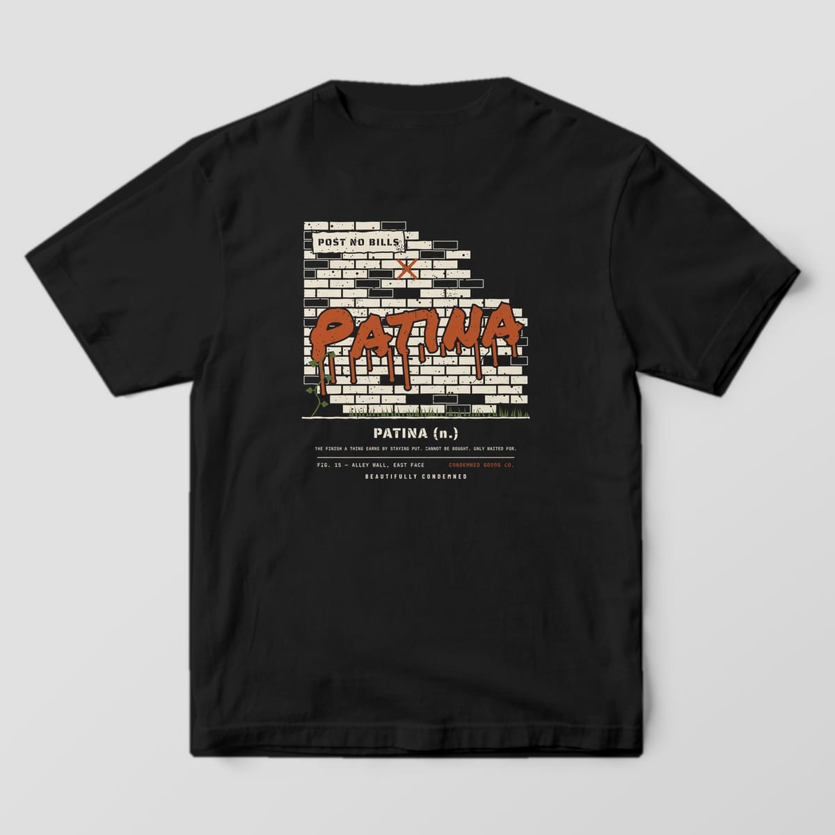 Back of a black Comfort Colors 1717 tee with a cream brick wall with the word Patina spray-painted in dripping rust.