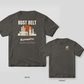Pepper tee shown back and front: back has a cream factory skyline with smokestacks against a striped rust sunset under the words Rust Belt Romantic; front has a small rusted gear with ferns.