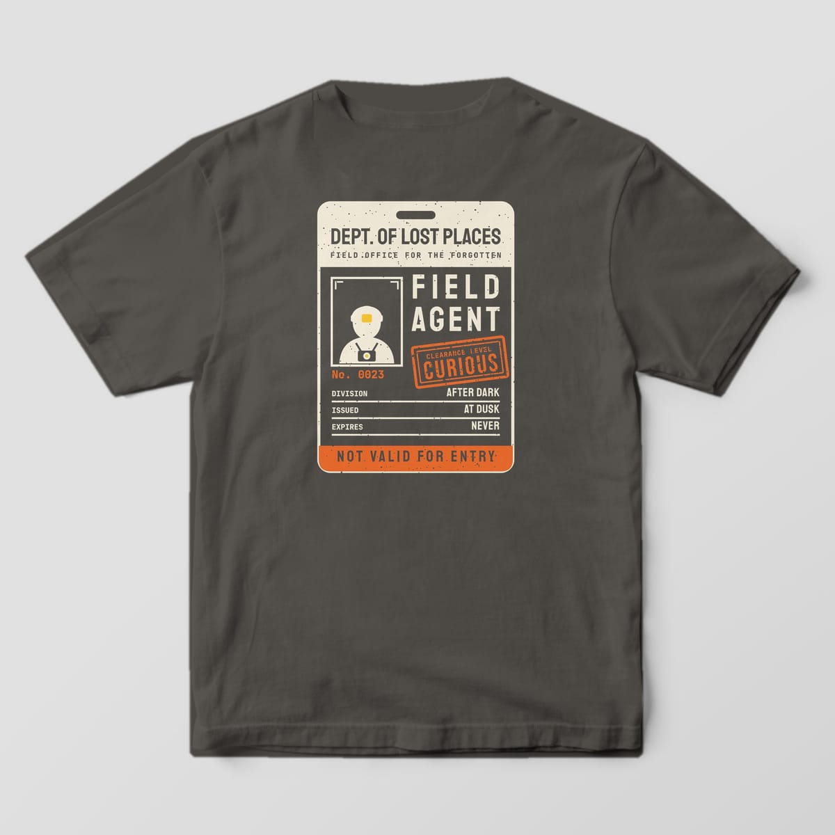 Back of a Pepper Comfort Colors 1717 tee printed with a cream, yellow and orange oversized ID card reading Dept. of Lost Places Field Agent with a headlamp explorer portrait and an orange Clearance Level Curious stamp.