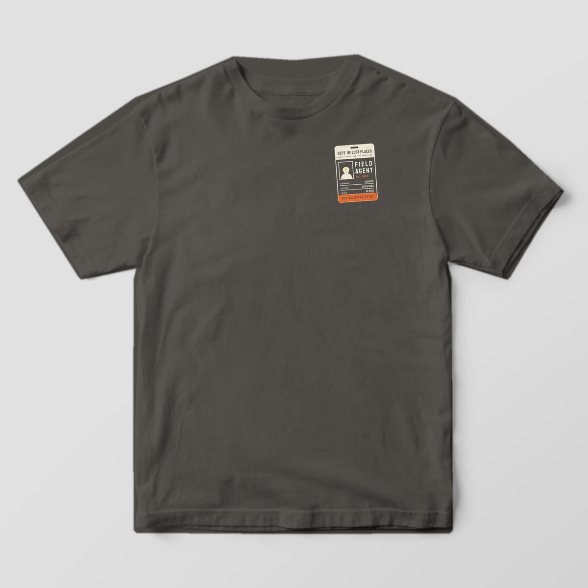 Front of a Pepper Comfort Colors 1717 tee with a small cream and orange Field Agent ID badge on the left chest.