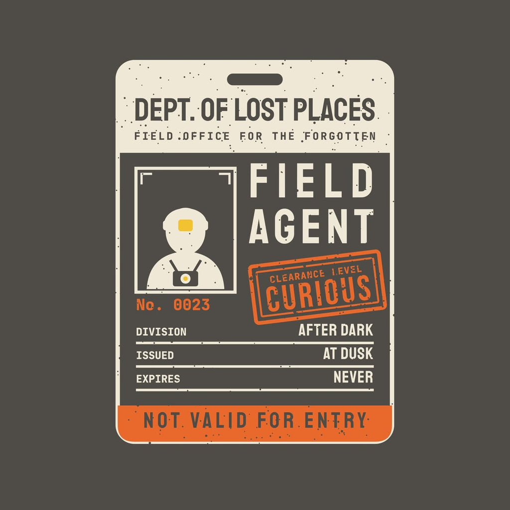 Back print artwork close-up: a cream, yellow and orange oversized ID card reading Dept. of Lost Places Field Agent with a headlamp explorer portrait and an orange Clearance Level Curious stamp, lightly speckled.