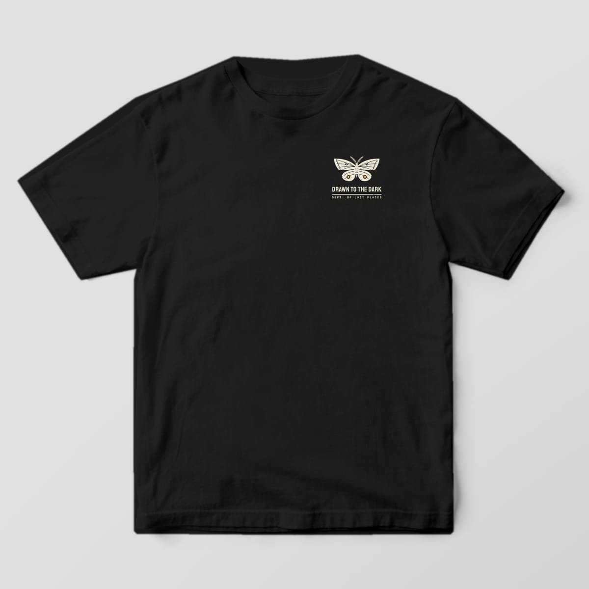 Front of a Black Comfort Colors 1717 tee with a small cream moth with yellow eyespots above the words Drawn to the Dark on the left chest.