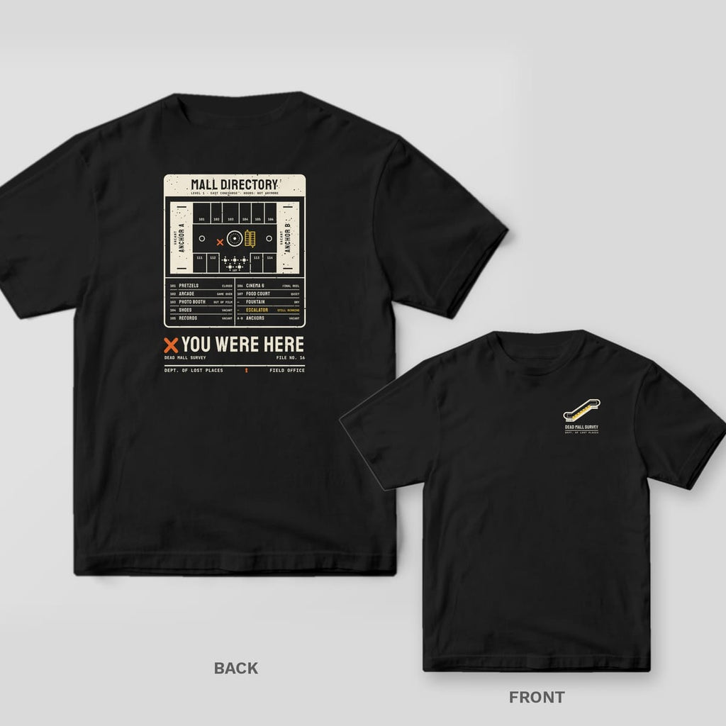 Black Comfort Colors tee shown back and front: the back has a cream dead mall directory map with an orange X above You Were Here; the chest has a small escalator.