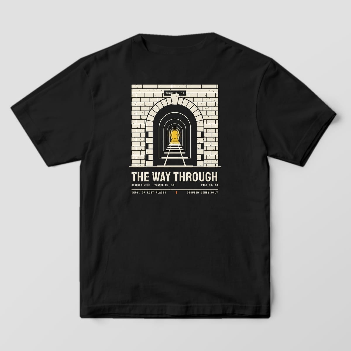 Back of a Black Comfort Colors 1717 tee printed with a cream brick railway tunnel portal with an arched entrance, rails running into the dark and a yellow light at the far end, above the words The Way Through.