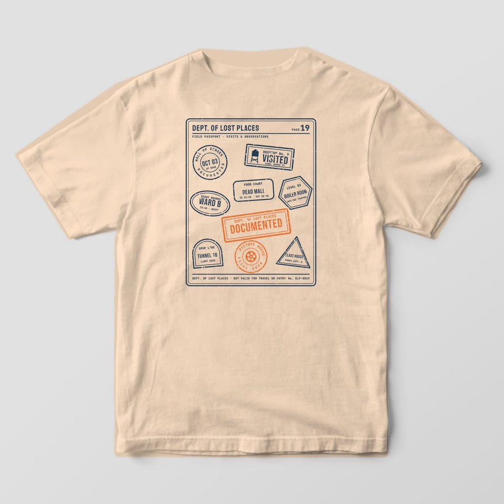 Back of an Ivory Comfort Colors 1717 tee printed with a navy passport-style page covered in rubber stamps from imaginary abandoned places, such as a rooftop, a tunnel and a dead mall, with an orange Documented stamp.