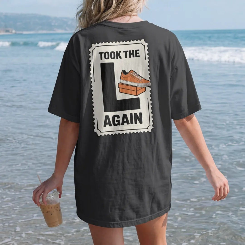 Woman on the beach in a Black tee, back view: a crumpled raffle ticket stamped with a giant L beside a sad shoebox, with Took The L above and Again below, as a two-ink streetwear graphic in off-white and orange.