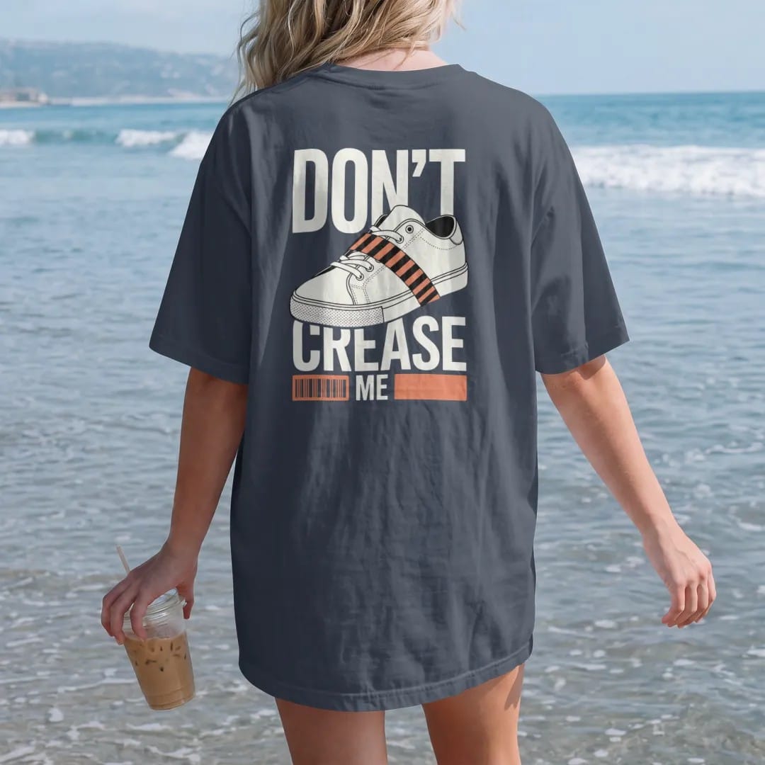 Woman on the beach in a Midnight tee, back view: a clean sneaker toe box wrapped in caution tape with Don't above and Crease Me below, as a two-ink streetwear graphic in off-white and orange.