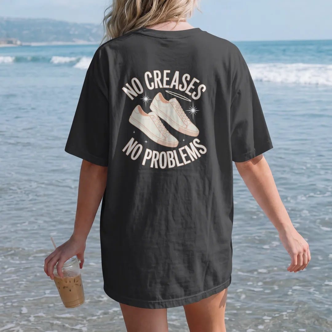 Woman on the beach in a Black tee, back view: a pair of spotless floating sneakers with sparkles and a halo glow, framed by No Creases and No Problems, as a two-ink streetwear graphic in off-white and orange.