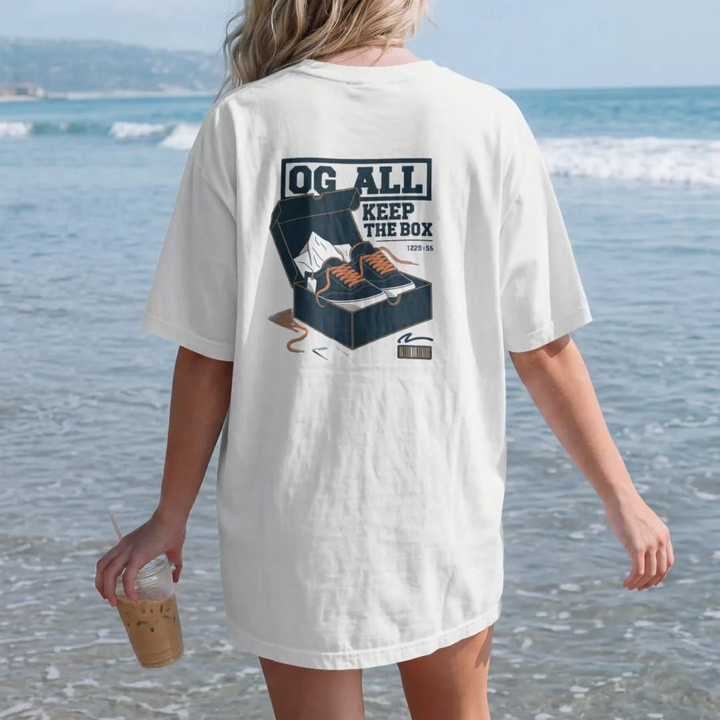 Woman on the beach in a White tee, back view: an open shoebox with tissue paper, spare laces and a hangtag under an OG All banner and Keep The Box script, as a two-ink streetwear graphic in navy and orange.