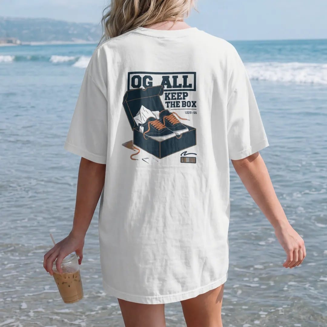 Woman on the beach in a White tee, back view: an open shoebox with tissue paper, spare laces and a hangtag under an OG All banner and Keep The Box script, as a two-ink streetwear graphic in navy and orange.