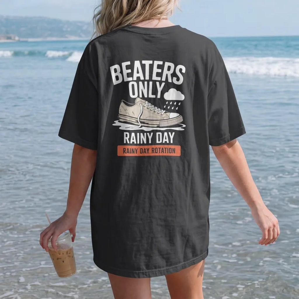 Woman on the beach in a Black tee, back view: a scuffed sneaker with a frayed lace splashing a puddle under a rain cloud, labeled Beaters Only and Rainy Day Rotation, as a two-ink streetwear graphic in off-white and orange.