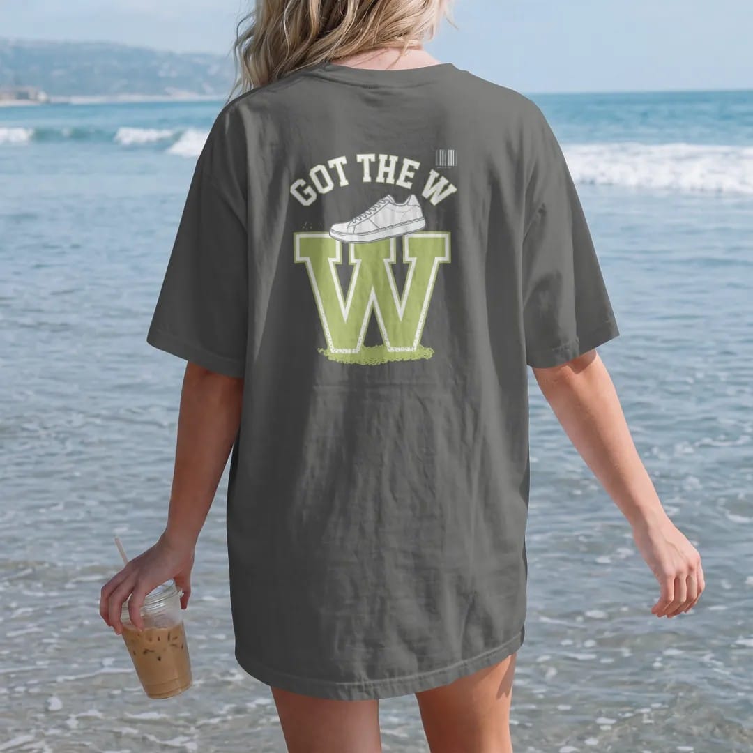 Woman on the beach in a Pepper tee, back view: a giant glowing W with a sneaker on top and bursting confetti under Got The W, as a two-ink streetwear graphic in off-white and lime.
