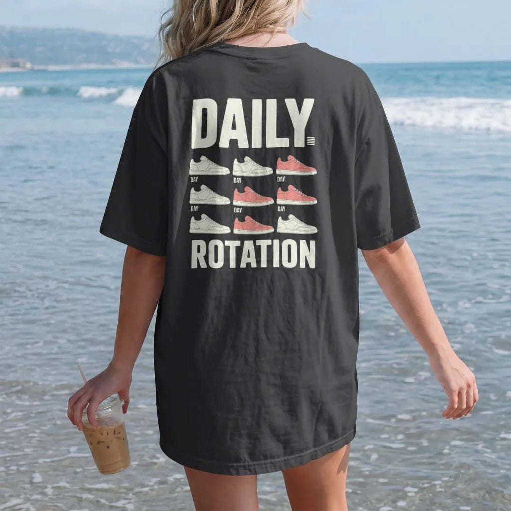 Woman on the beach in a Black tee, back view: a weekly planner grid of seven different sneakers under Daily and above a big Rotation, as a two-ink streetwear graphic in off-white and orange.