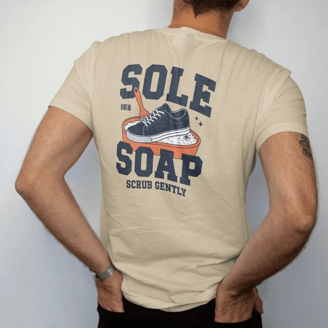 Man wearing a Sandstone tee, seen from the back: a retro ad of a smiling sneaker scrubbing itself in a sudsy tub with Sole Soap in bubble letters and Scrub Gently below, as a two-ink streetwear graphic in navy and orange.