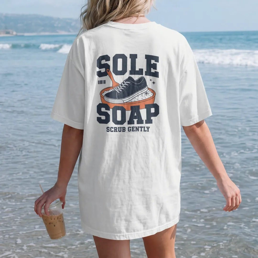 Woman on the beach in a White tee, back view: a retro ad of a smiling sneaker scrubbing itself in a sudsy tub with Sole Soap in bubble letters and Scrub Gently below, as a two-ink streetwear graphic in navy and orange.