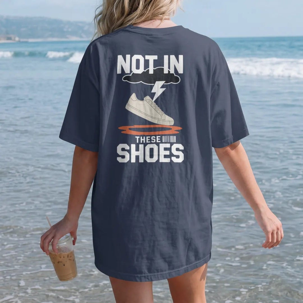Woman on the beach in a Navy tee, back view: a sneaker leaping over a puddle under a rain cloud with Not In above and These Shoes below, as a two-ink streetwear graphic in off-white and orange.