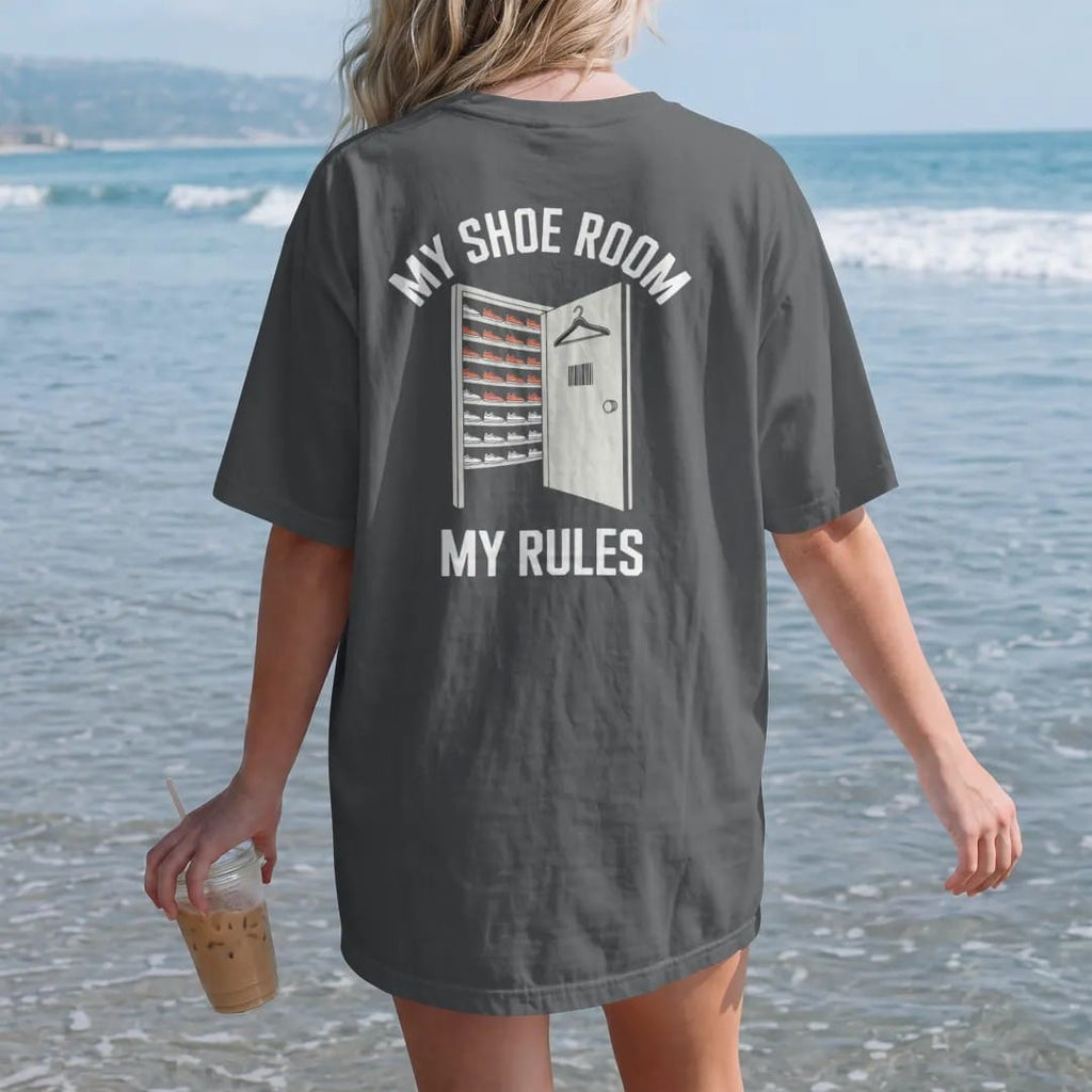 Woman on the beach in a Graphite tee, back view: a doorway into a room of lit sneaker shelves with a door hanger, under My Shoe Room and My Rules, as a two-ink streetwear graphic in off-white and orange.