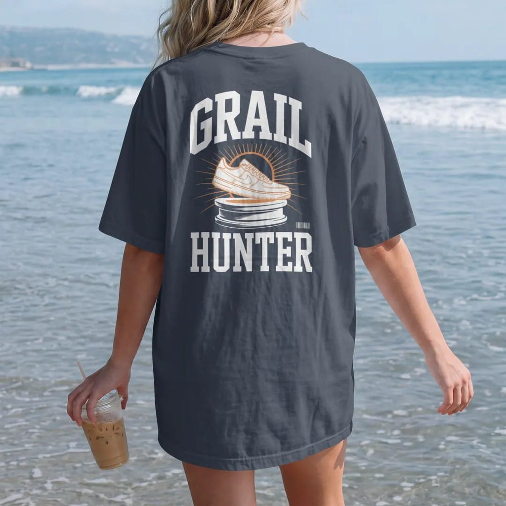 Woman on the beach in a Midnight tee, back view: a sneaker glowing on a stone pedestal with a halo and light rays, with Grail arched above and Hunter below, as a two-ink streetwear graphic in off-white and orange.