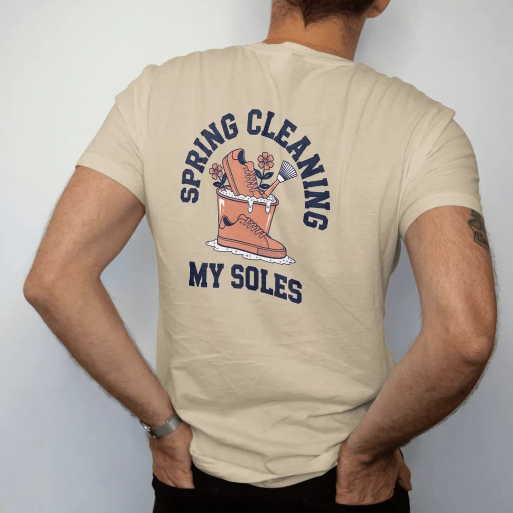 Man wearing a Sandstone tee, seen from the back: a sneaker in a sudsy bucket with blooming flowers and a scrub brush, under Spring Cleaning and above My Soles, as a two-ink streetwear graphic in navy and orange.