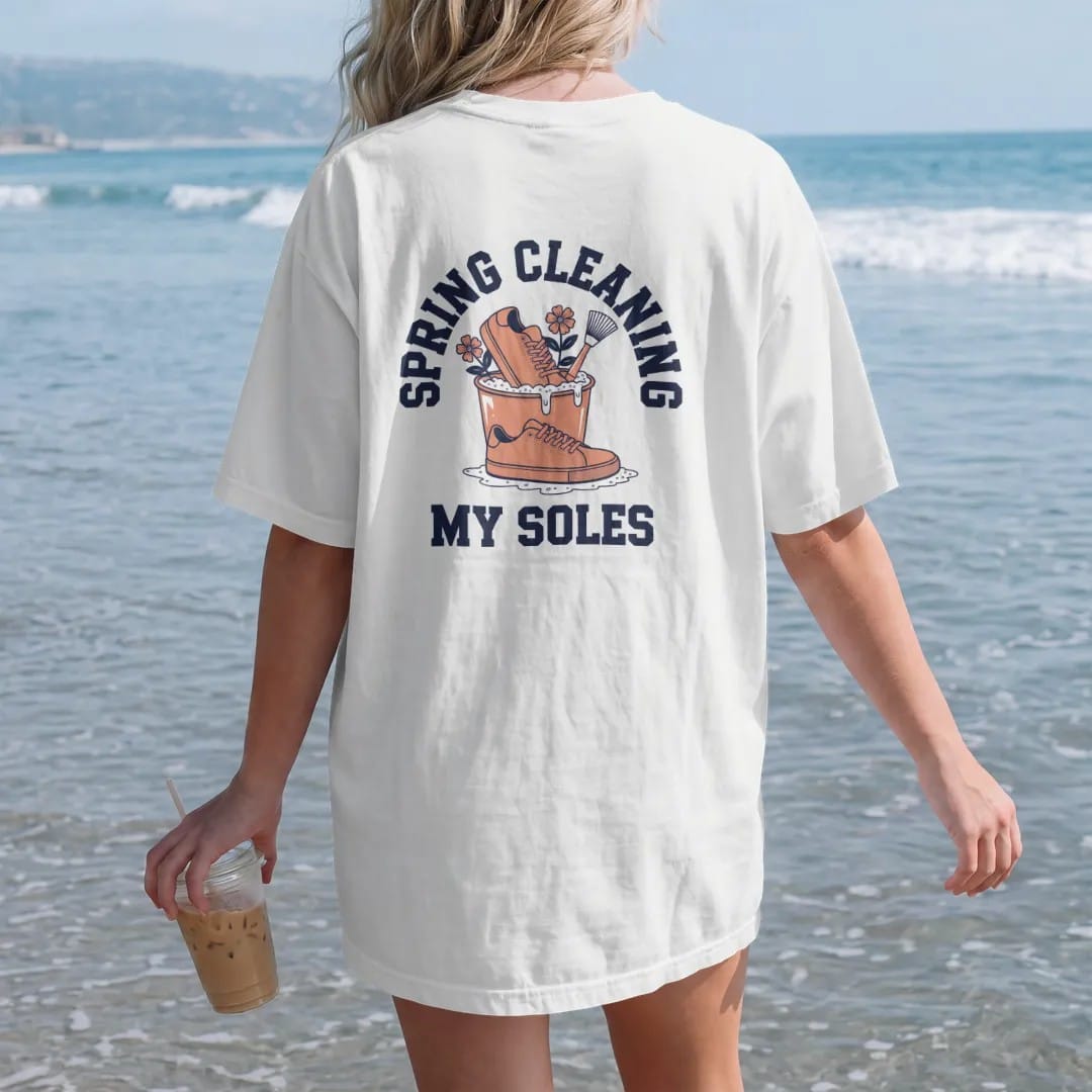Woman on the beach in a White tee, back view: a sneaker in a sudsy bucket with blooming flowers and a scrub brush, under Spring Cleaning and above My Soles, as a two-ink streetwear graphic in navy and orange.
