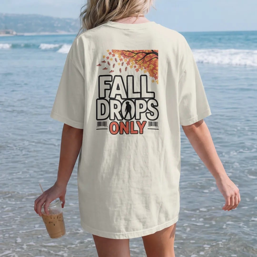 Woman on the beach in a Ivory tee, back view: autumn leaves and tiny sneakers falling from a tree with Fall Drops arched above and Only below, as a two-ink streetwear graphic in off-white and orange.