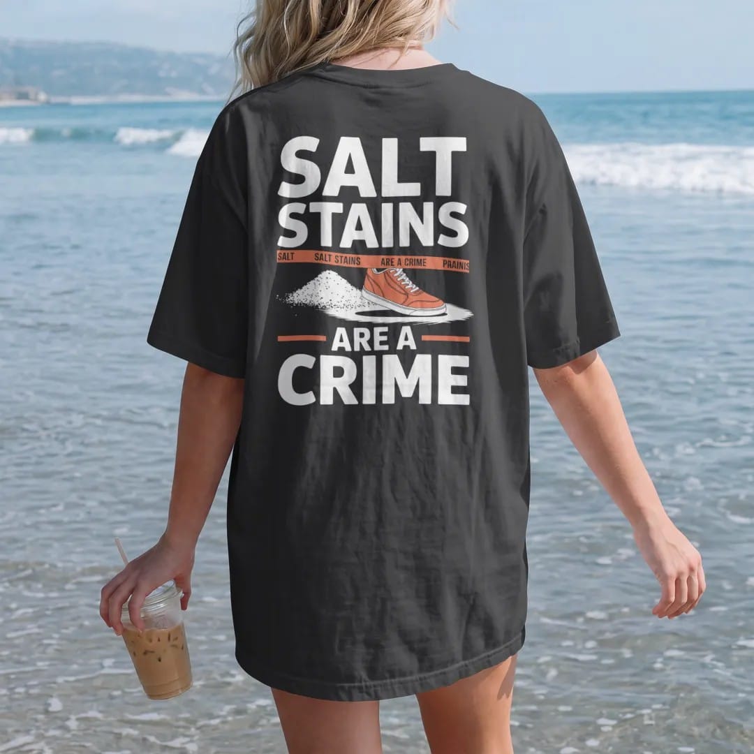 Woman on the beach in a Black tee, back view: a sneaker tiptoeing around road salt on a snowy sidewalk with police tape reading Salt Stains and Are A Crime below, as a two-ink streetwear graphic in off-white and orange.