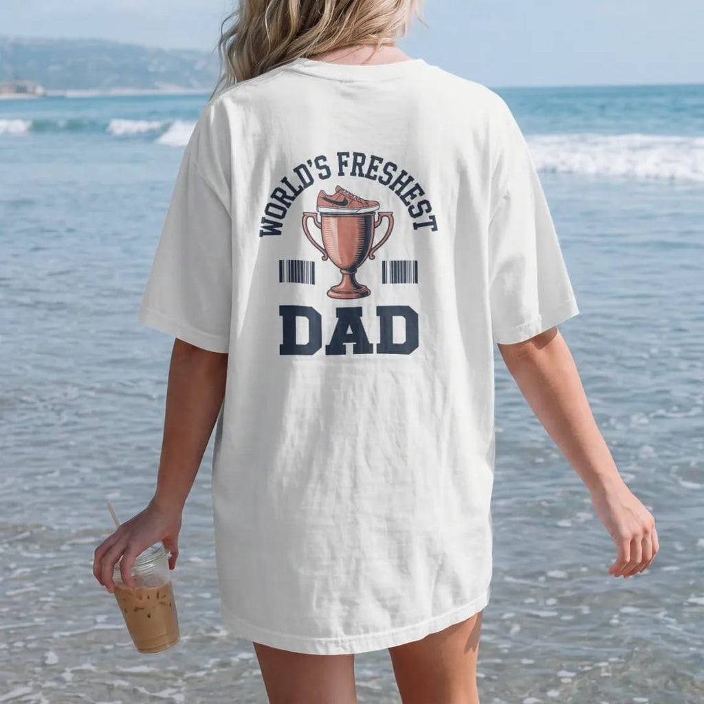 Woman on the beach in a White tee, back view: a trophy cup topped with a sneaker and a ribbon under World's Freshest and a giant Dad, as a two-ink streetwear graphic in navy and orange.
