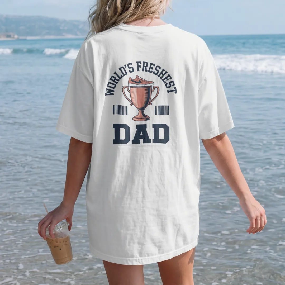 Woman on the beach in a White tee, back view: a trophy cup topped with a sneaker and a ribbon under World's Freshest and a giant Dad, as a two-ink streetwear graphic in navy and orange.