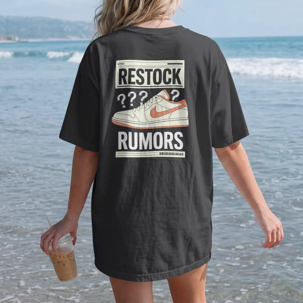 Woman on the beach in a Black tee, back view: a vintage newspaper page with a sneaker photo and scribbled arrows, masthead Restock and headline Rumors, as a two-ink streetwear graphic in off-white and orange.
