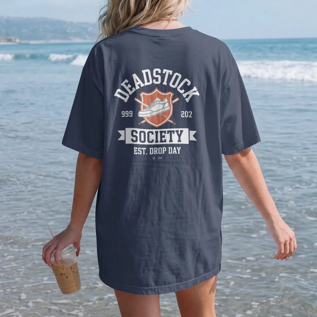 Woman on the beach in a Navy tee, back view: a varsity shield with a sneaker and crossed laces, Deadstock arched above, Society script banner and Est. Drop Day below, as a two-ink streetwear graphic in off-white and orange.